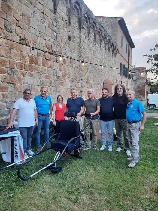 Gruppo di persone in posa intorno a carrozzina joelette. Muro sullo sfondo.