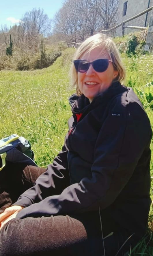 Donna seduta per terra, verde e alberi sullo sfondo.